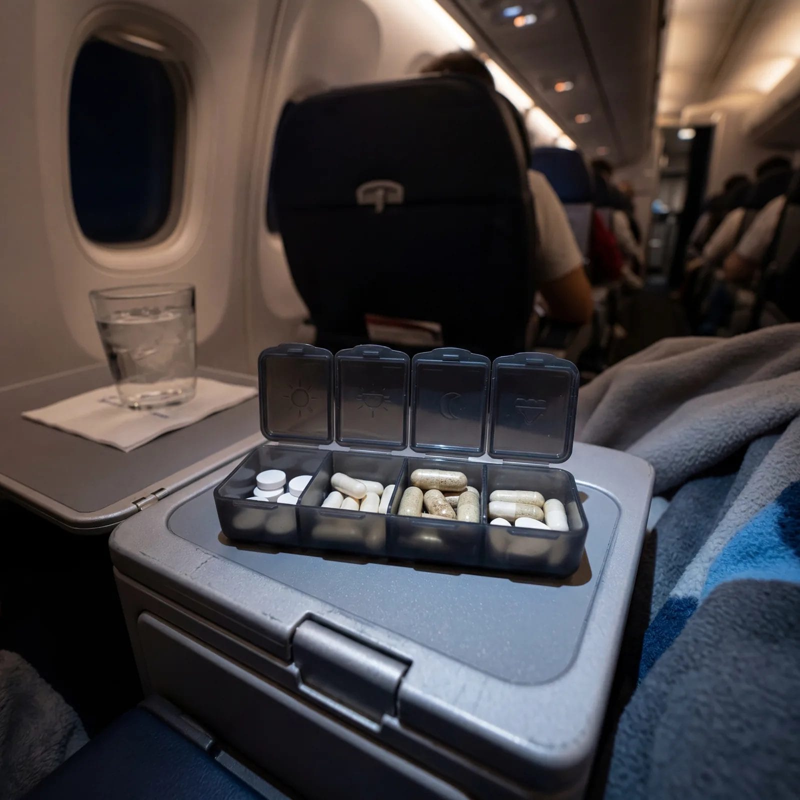 Small travel pill organizer with melatonin tablets and sleep support supplements on an airplane tray table, cabin lighting, photorealistic, editorial travel photography, no text, no watermark, 16:9